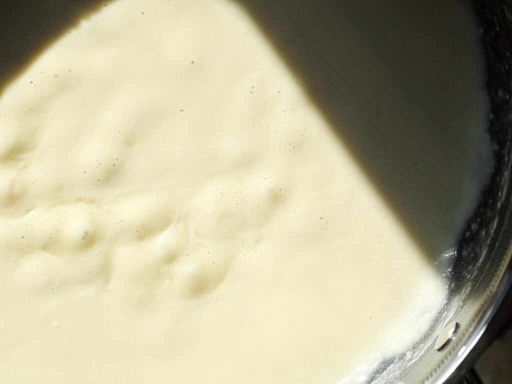 Close-up of creamy sauce in a metallic mixing bowl partially covered by a shadow. Small bubbles dot the smooth surface, capturing the mix of light and shadow across the batter.